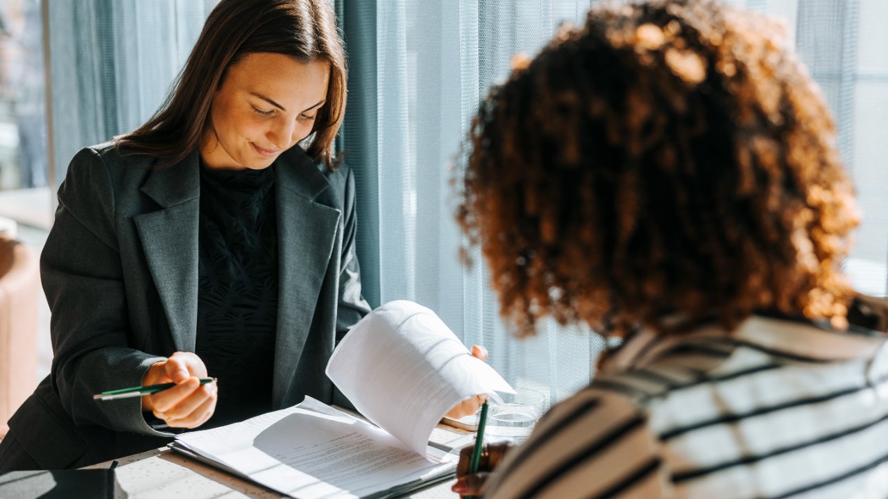 Two women sit across from each other at a table while reviewing paperwork.