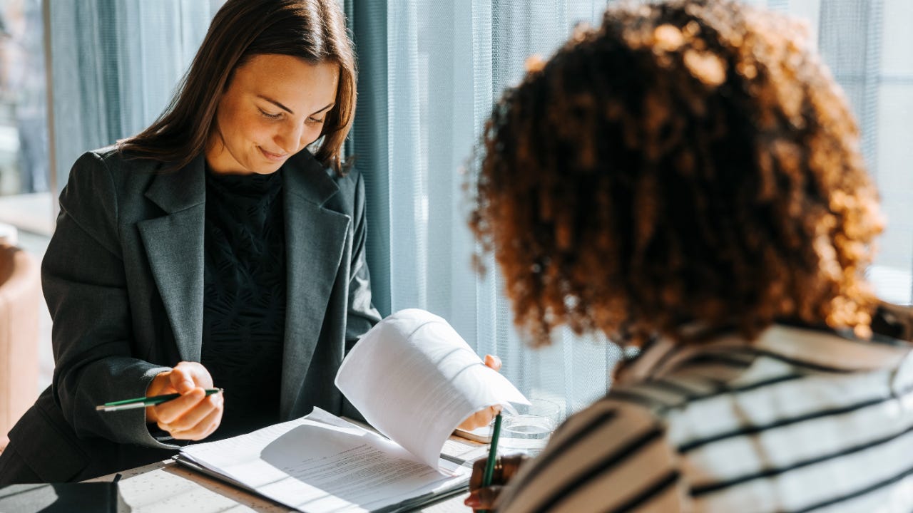 Two women sit across from each other at a table while reviewing paperwork.