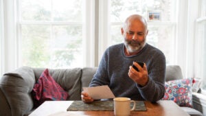 A man sits on a couch while reading his phone and reviewing a piece of mail.