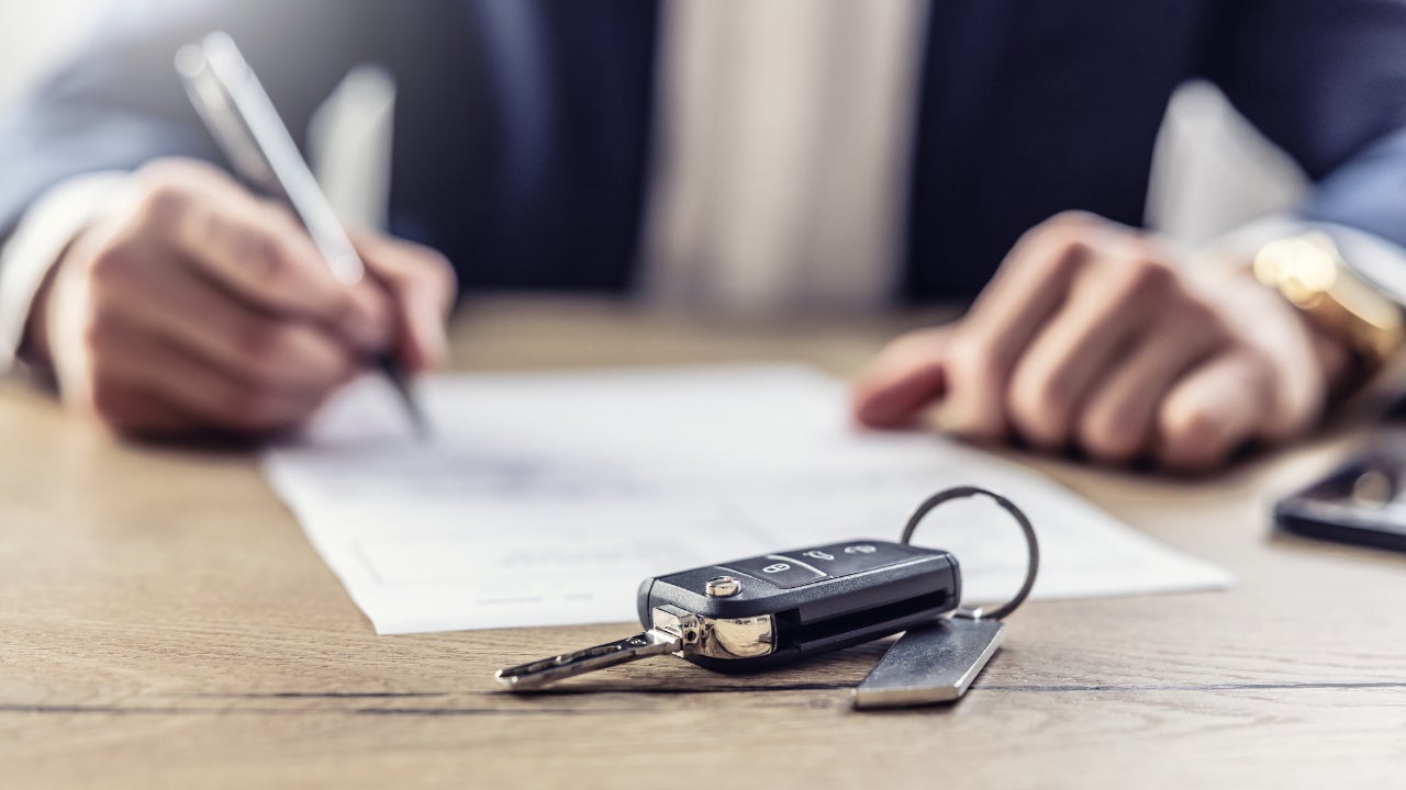 A set of hands signing a document. In the foreground, there is a set of car keys.