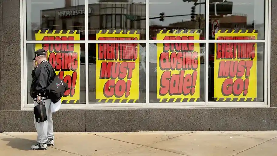 A man standing outside a store with signs in the window proclaiming 'Everything Must Go.'